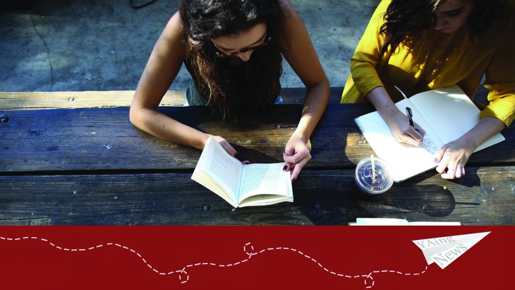 Two people sit at a park bench in the sunshine: one writing in a notebook, and one reading a book. 