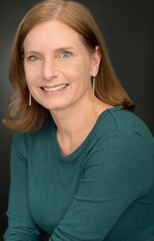 Tanya Lloyd Kyi sits facing left with her head turned to face the camera, smiling. She wears long earrings and a dark longsleeve shirt.