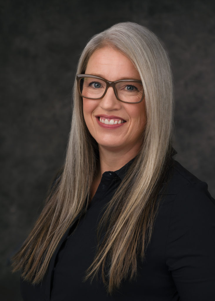 Susan Juby smiles in her author photo. She sports dark square glasses, a black shirt, and long, ombre grey hair.