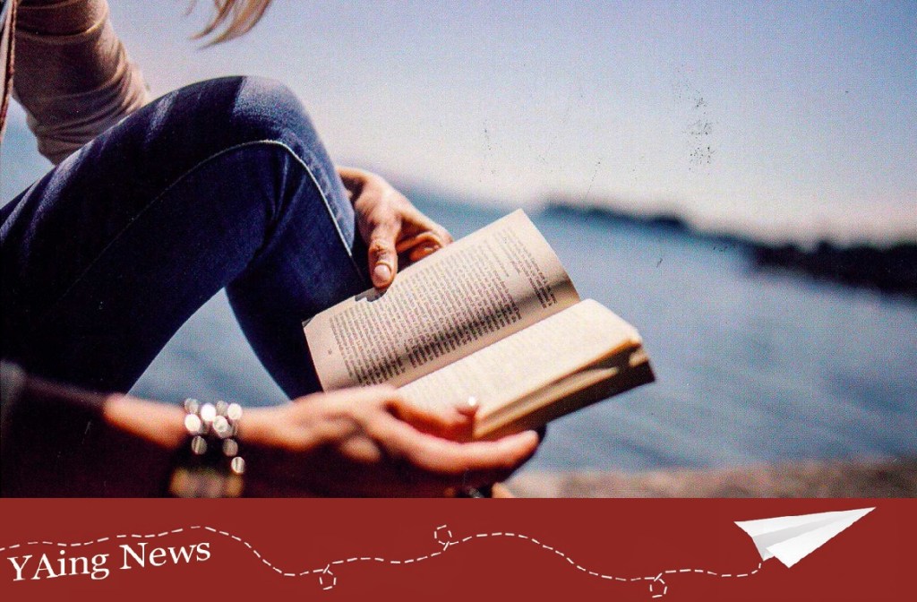 A person reads a book on the beach in sunlight. The red YAing News tag is beneath.